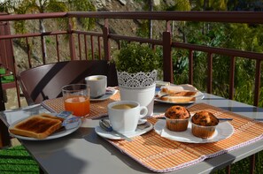 Una mesa servida con alimentos para el desayuno, incluyendo tostadas, muffins, una taza de café y un vaso de jugo de naranja, con vista a follaje en el fondo.