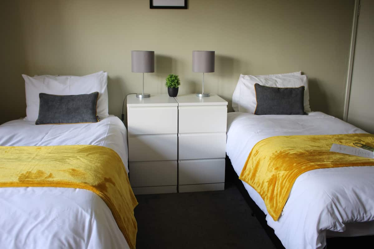 Quarto de hotel com duas camas de solteiro, cada uma com um edredom branco e um toque de cor amarelo. Há duas mesas de cabeceira com luminárias e uma pequena planta em vaso.