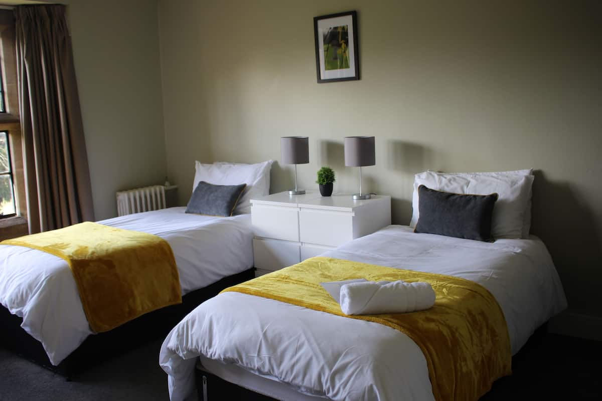 Quarto de hotel com duas camas, cada uma com colcha branca e manta amarela. Há duas luminárias de cabeceira e um pequeno vaso de planta sobre a mesa de cabeceira branca entre as camas.