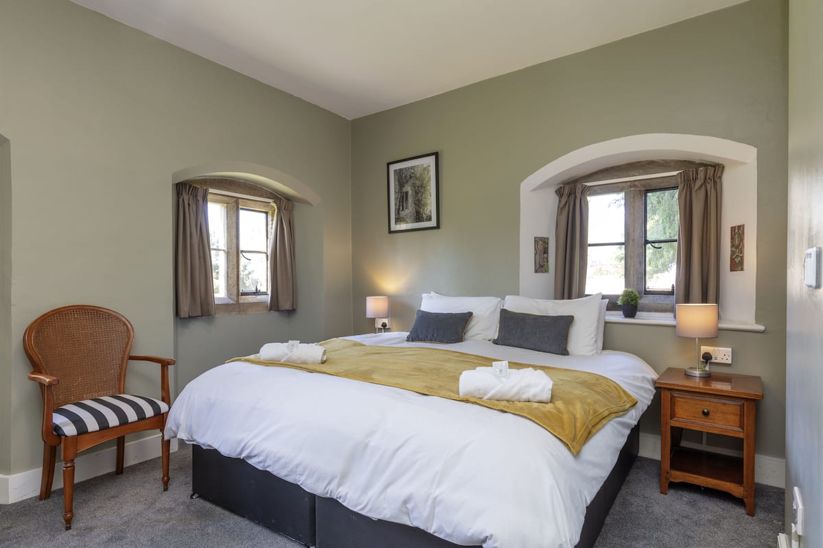Uma cama bem arrumada com roupas de cama brancas e douradas, uma cadeira de madeira com almofadas listradas e duas mesas de cabeceira com abajures.
