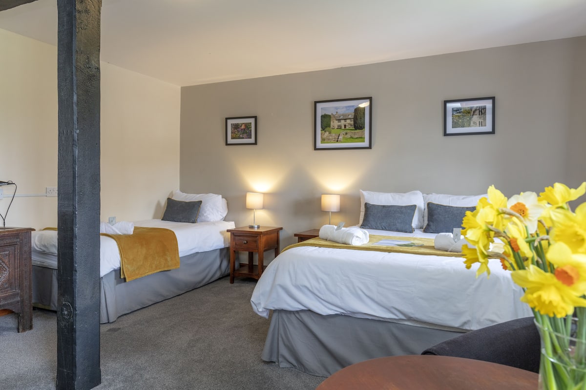 Quarto de hotel com duas camas, uma mesa de cabeceira de madeira, uma pequena mesa e um vaso com flores amarelas.