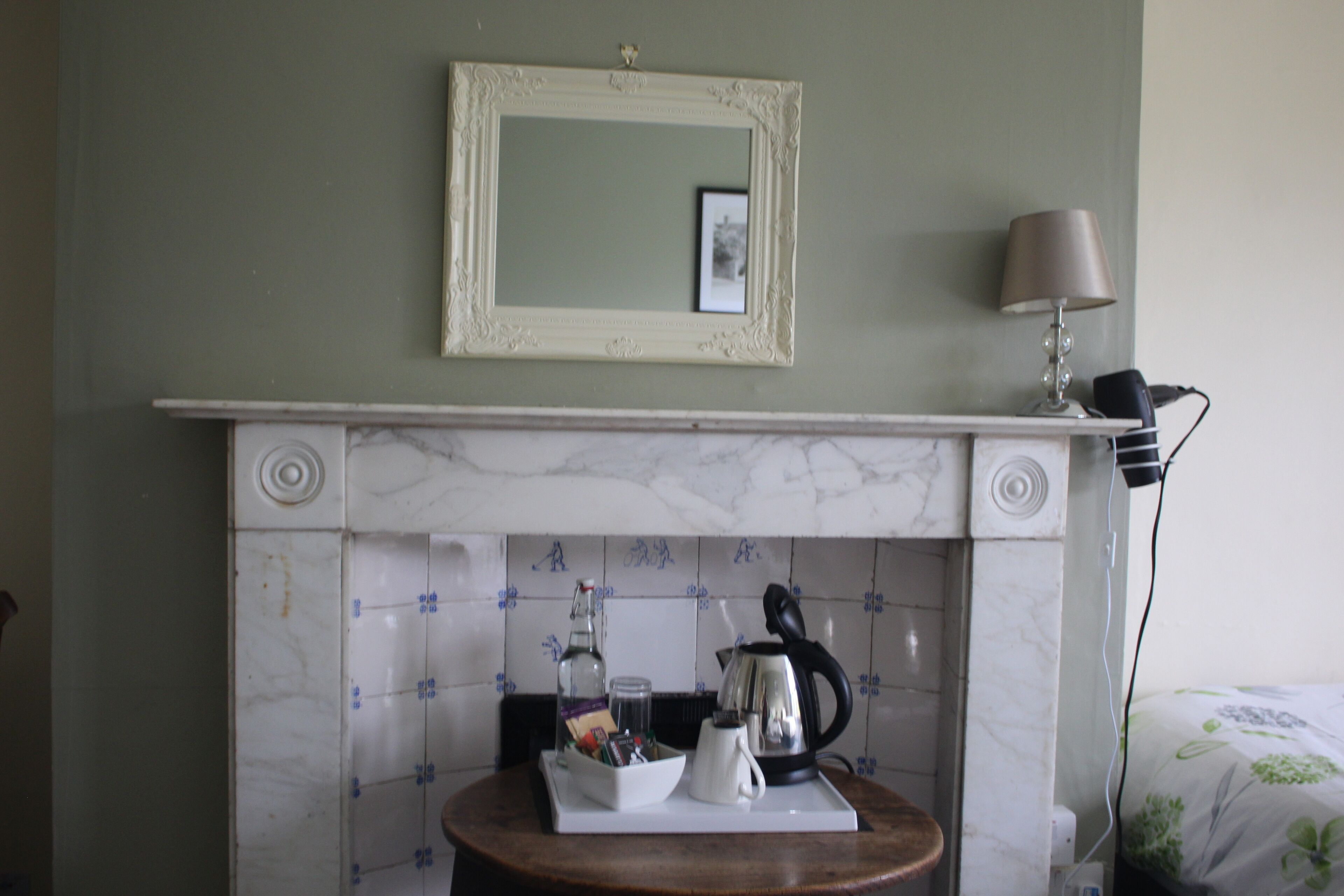 A room with a fireplace, a framed mirror, a lamp, and a table with a kettle and a tray.
