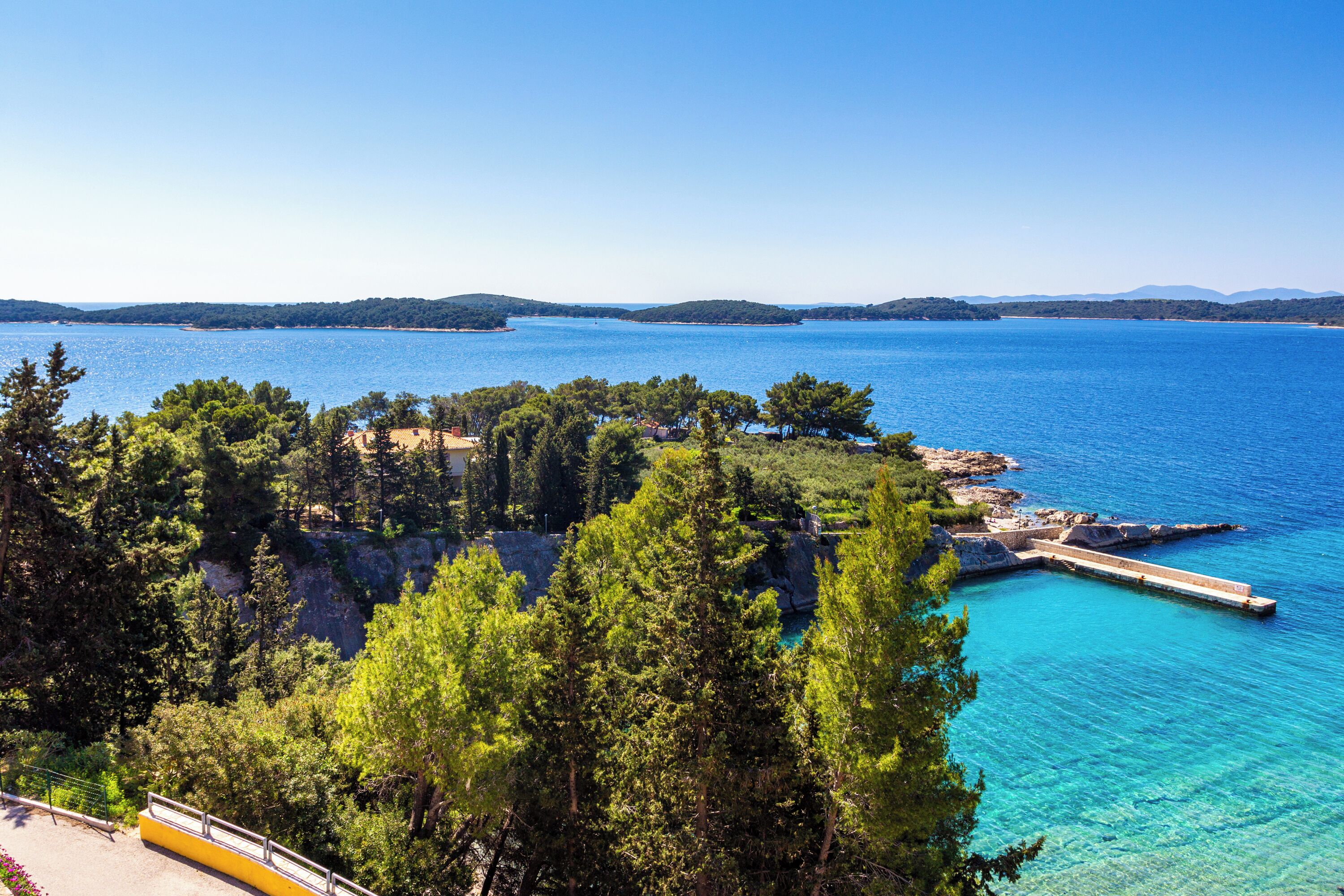 A coastal landscape with a pier extending into the sea, surrounded by dense greenery and clear blue waters.