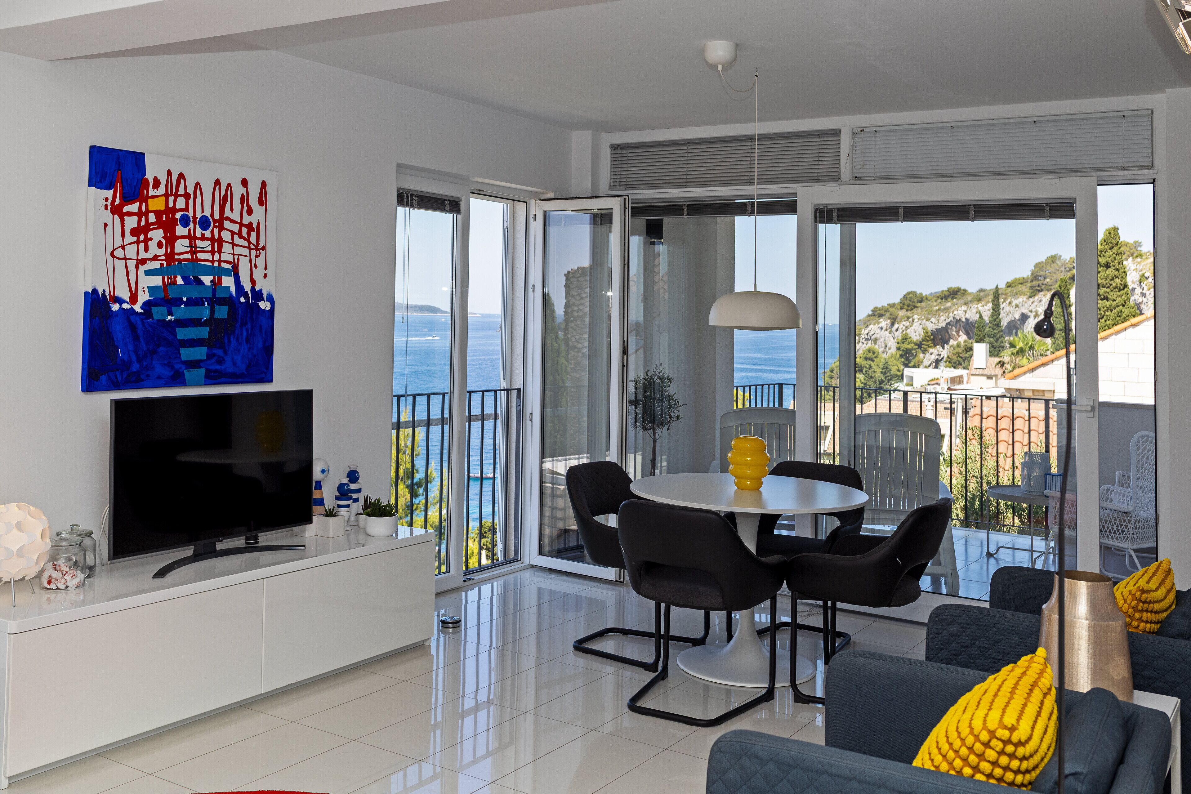 A modern living room with a flat-screen TV, a dining area with a round table and chairs, and a balcony with a view of the sea.