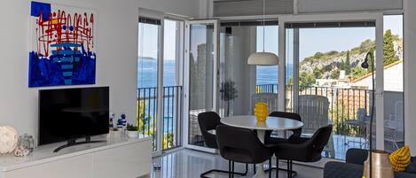A modern living room with a flat-screen TV, a dining area with a round table and chairs, and a balcony with a view of the sea.