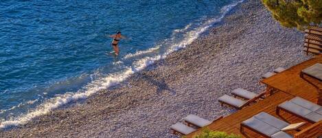 A beach with a person walking in the water, a wooden deck with lounge chairs, and a rocky shoreline.