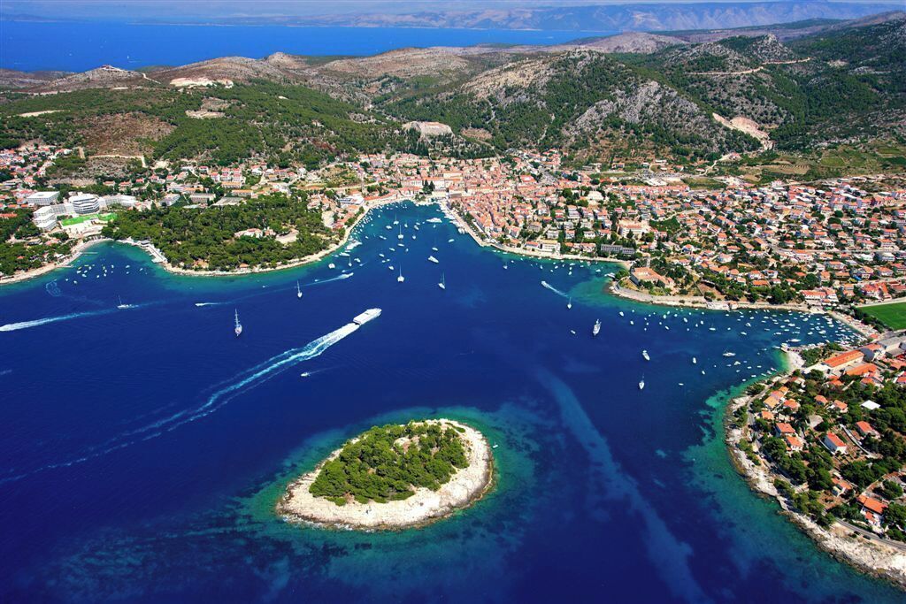 Vista aérea de un pueblo costero con un puerto deportivo, una pequeña isla y colinas circundantes.