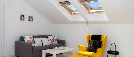 Un salon moderne avec un canapé gris, un fauteuil jaune, une table basse blanche et une lucarne au plafond.