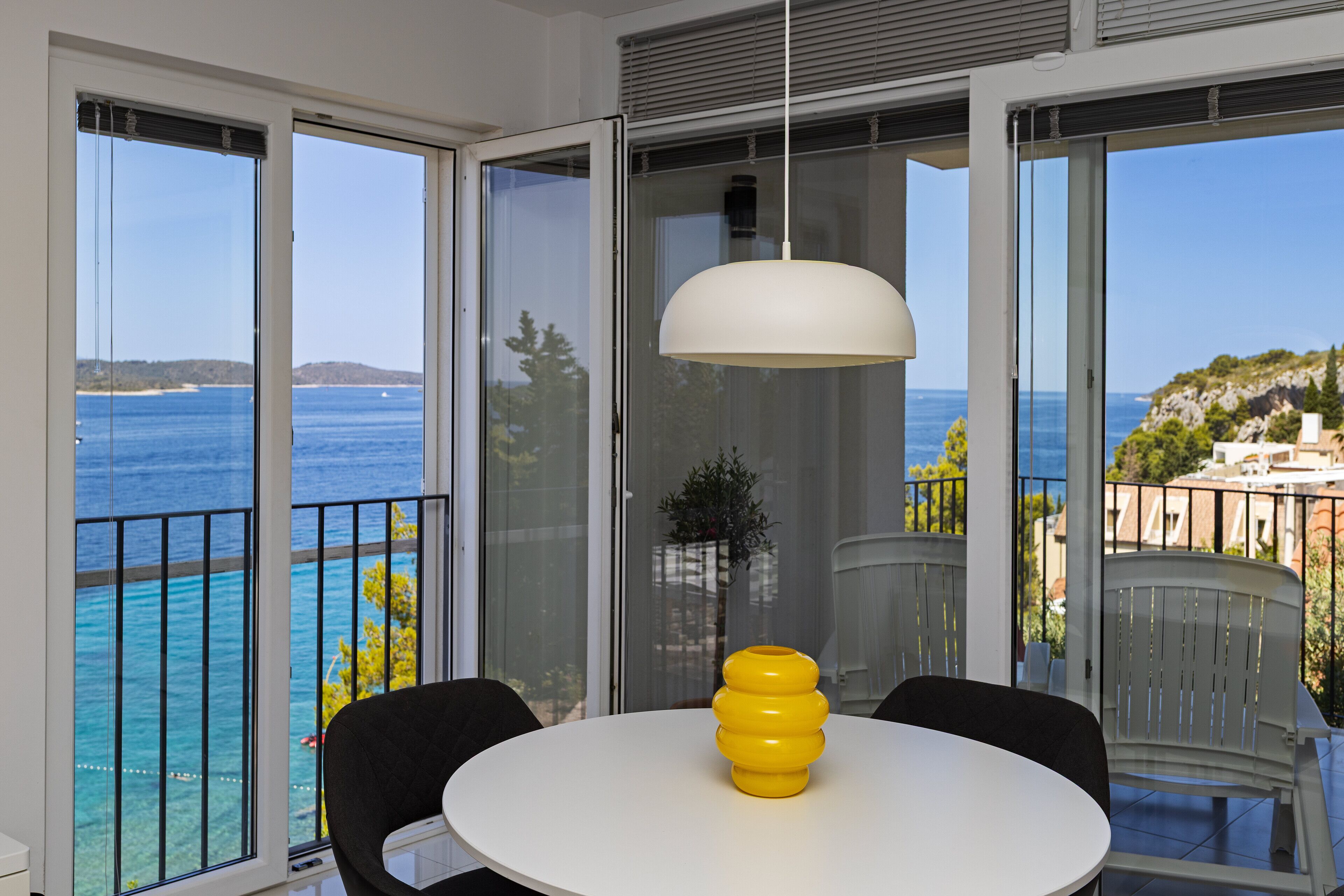 Un balcón con vista al mar y una mesa blanca con un jarrón amarillo.