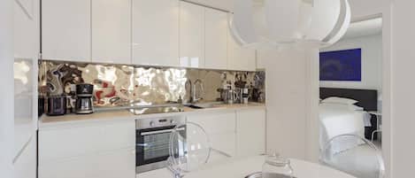 A modern kitchen with white cabinets, a stainless steel oven, and a coffee maker. There is a dining table with a glass dome cover and a tray.
