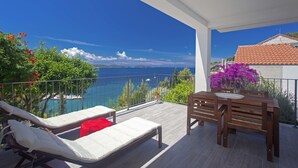 A balcony with a wooden table and two lounge chairs, overlooking a clear blue sea and a mountainous landscape. - Blue Bay Residence (Hvar)