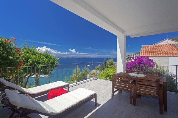 Un balcón con una mesa de madera y dos sillones de lounge, con vista a un mar azul y un paisaje montañoso.