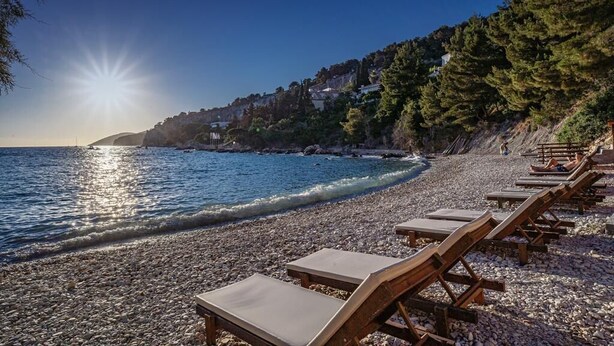A pebble beach with sunlit water, wooden lounge chairs, and a hillside with buildings. - Blue Bay Residence (Hvar)