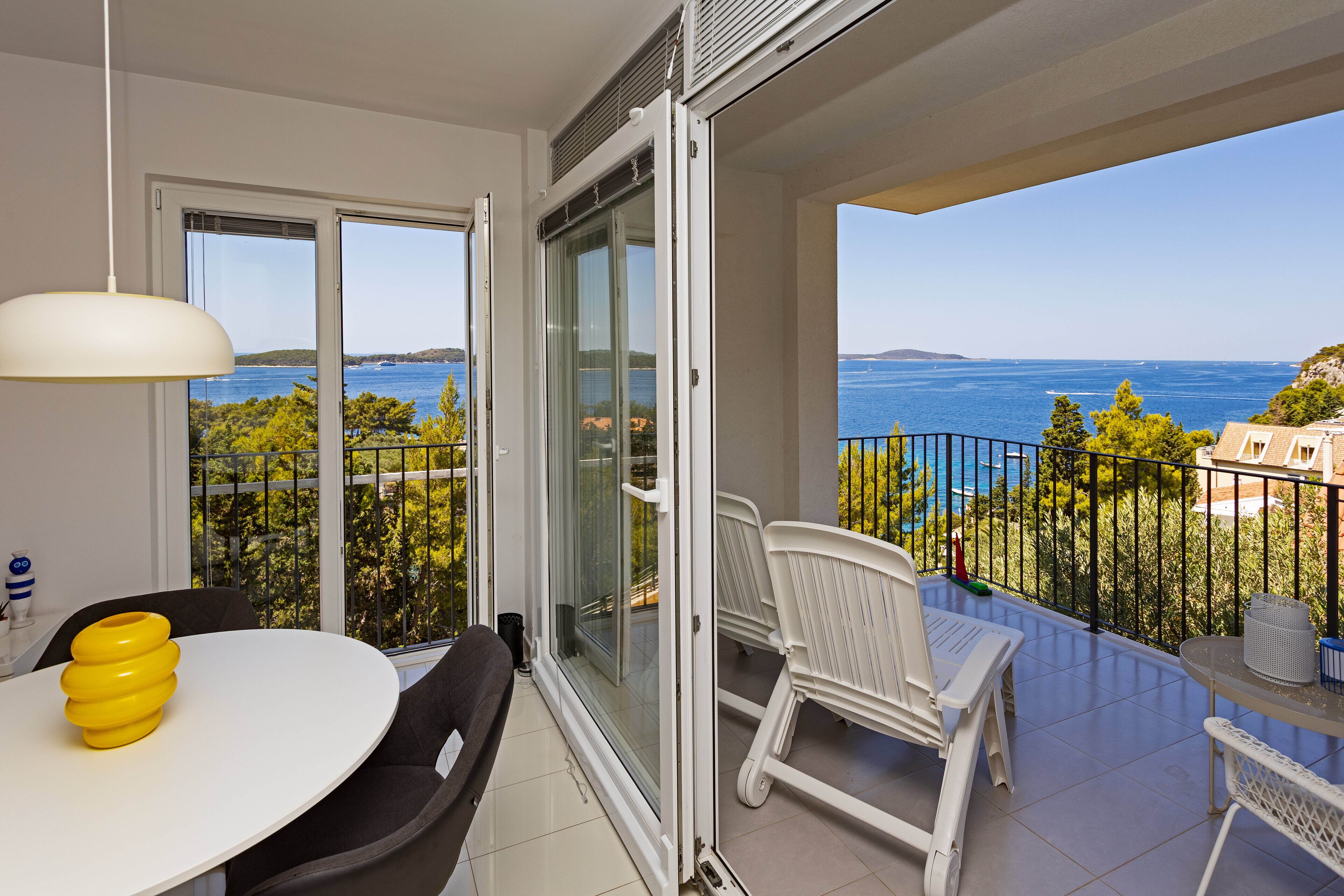 Un balcon avec vue sur la mer, un coin repas avec une table et des chaises blanches, et un luminaire suspendu blanc.