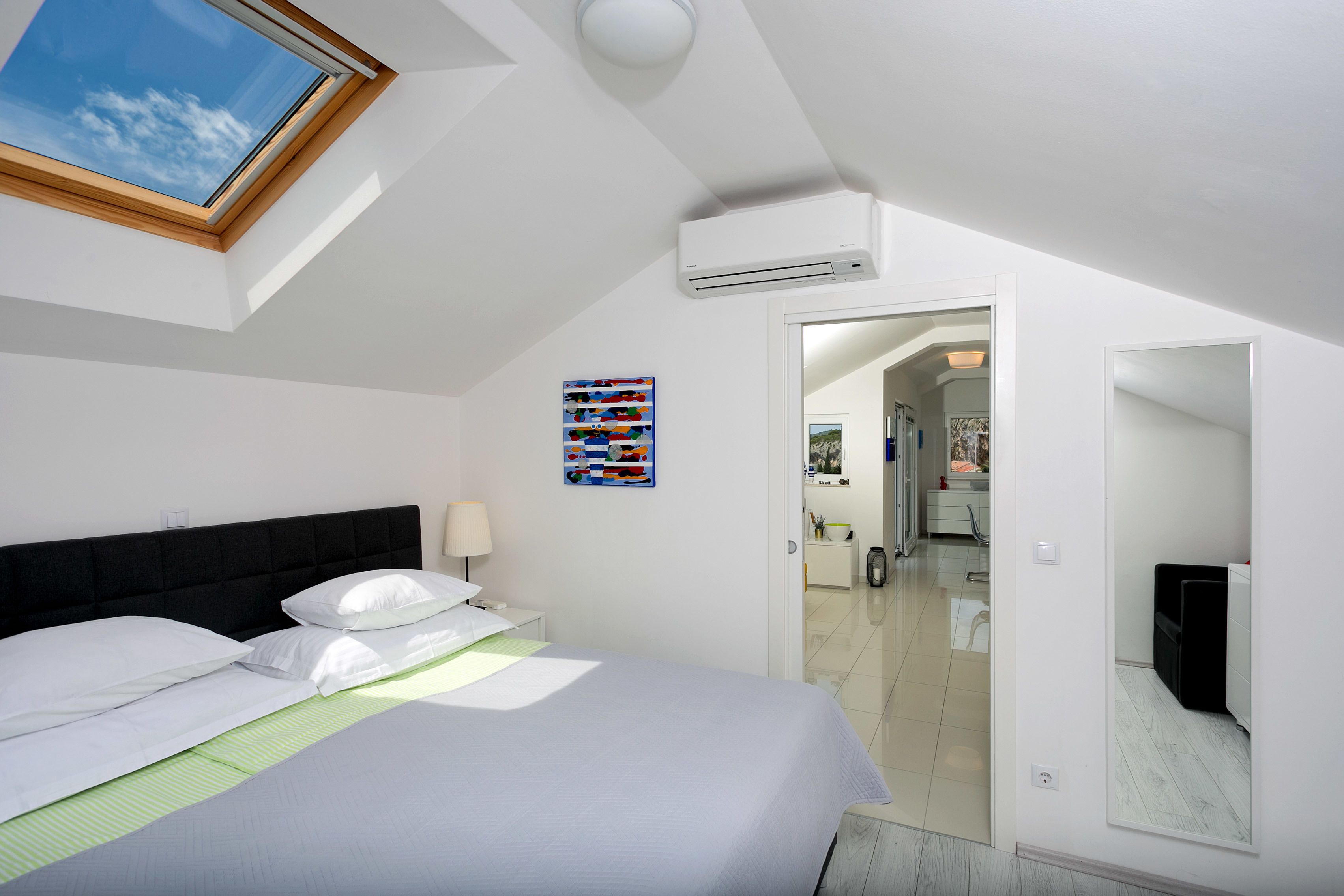 A bedroom with a skylight, a bed with white bedding, a bedside table with a lamp, and a colorful artwork on the wall.