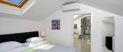 A bedroom with a skylight, a bed with white bedding, a bedside table with a lamp, and a colorful artwork on the wall.