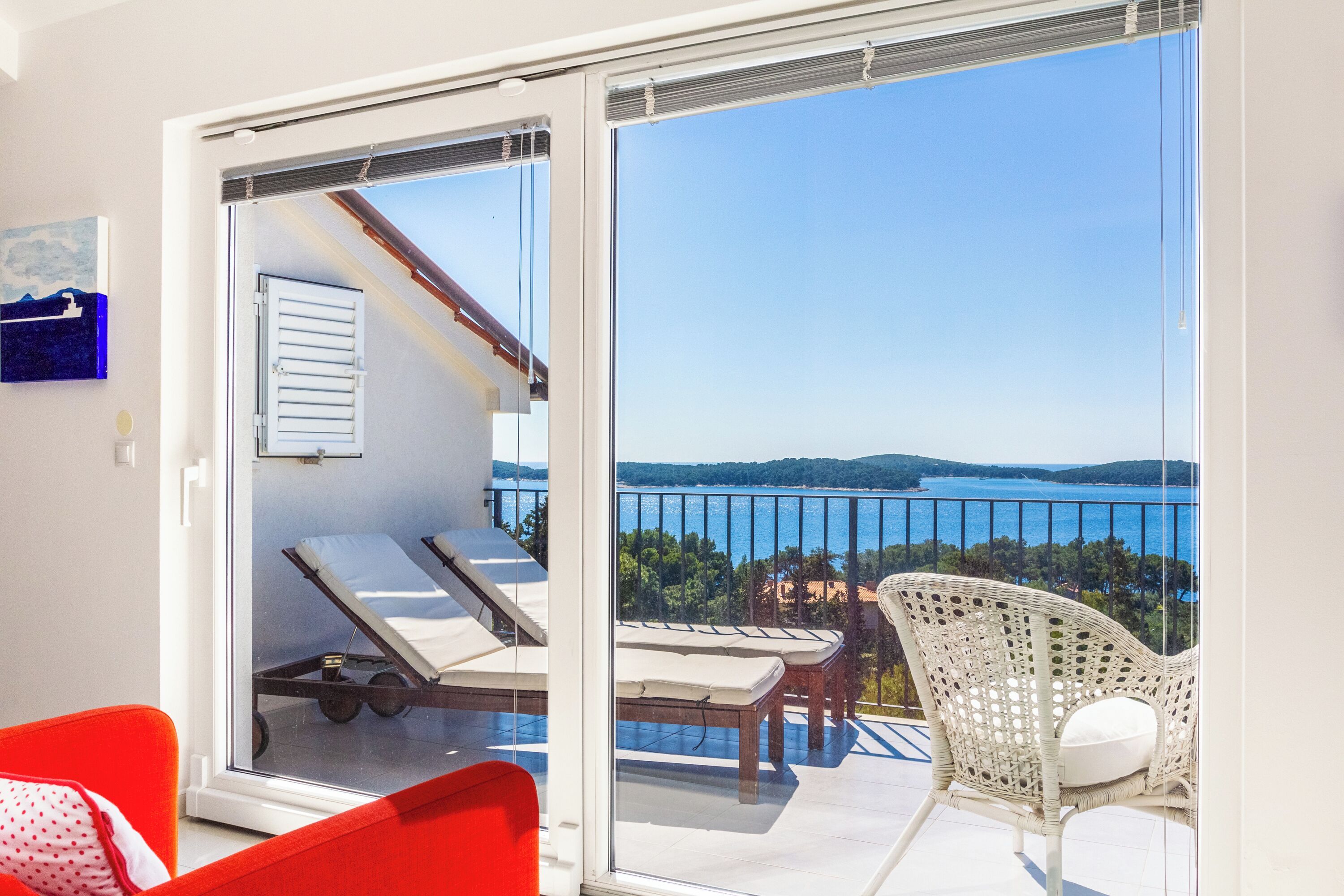 A balcony with a view of a lake and hills, a white wicker chair, and a red chair.
