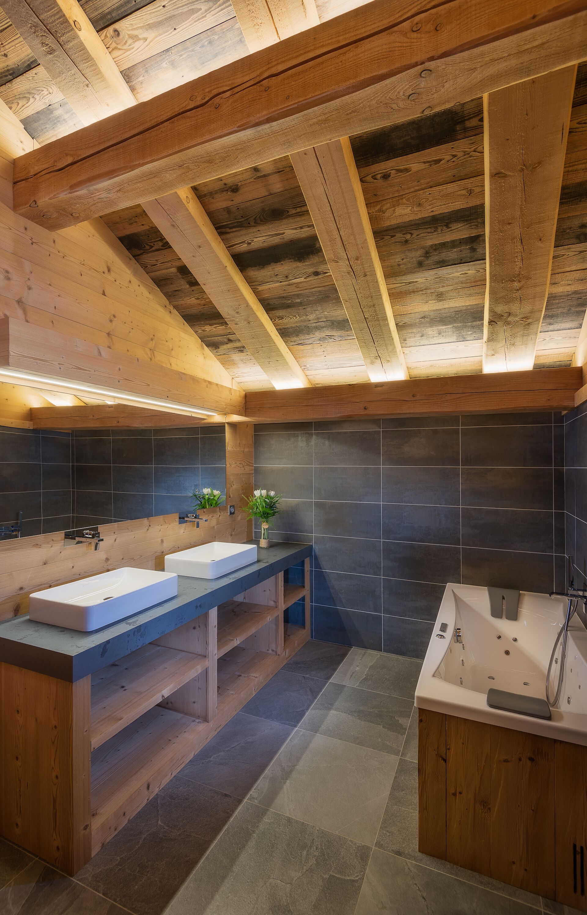 Un bagno moderno con travi in legno, un ampio specchio, due lavandini, una vasca da bagno e pareti piastrellate di colore scuro.