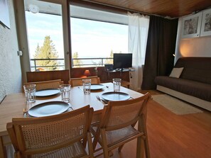 Ruokailutila, jossa on neljälle katettu ruokapöytä, puiset tuolit, sohva, televisio ja näkymä puille ikkunasta.