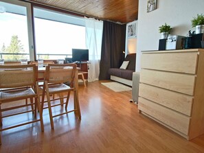 A modern living room with wooden furniture, a sofa, a coffee table, and a dining area with chairs. There is a large window providing natural light and a view of the outdoors.