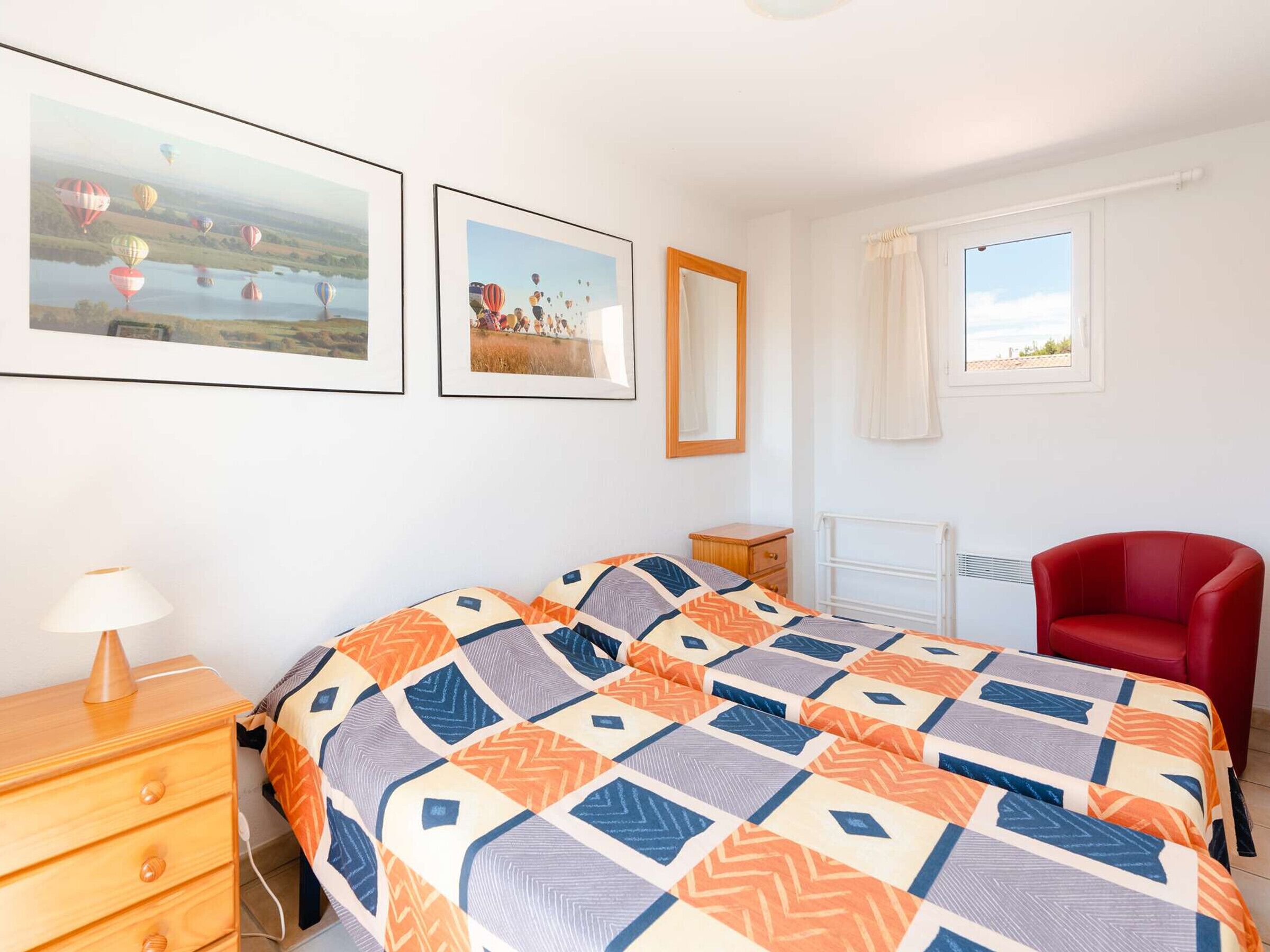 Ein Schlafzimmer mit einem Bett, einem Nachttisch, einer Lampe, einem Sessel, einem Fenster und zwei gerahmten Bildern an der Wand.