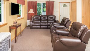 A living room with a brown leather sofa, a flat-screen TV, a ceiling fan, and a window with red curtains.