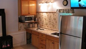 A compact kitchen with wooden cabinets, a microwave, a toaster, a sink, a refrigerator, and a wall-mounted clock.