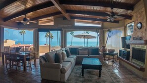 Amplia sala de estar con un sofá grande, una mesa de centro y un comedor. Un televisor está montado en la pared, y hay una chimenea. La habitación tiene ventanas amplias que ofrecen vistas a la playa y al océano.