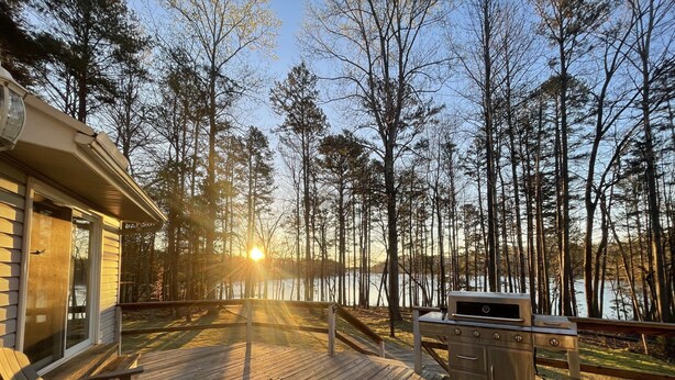 Une terrasse en bois avec des fauteuils Adirondack, un barbecue et une vue sur un lac entouré d’arbres.