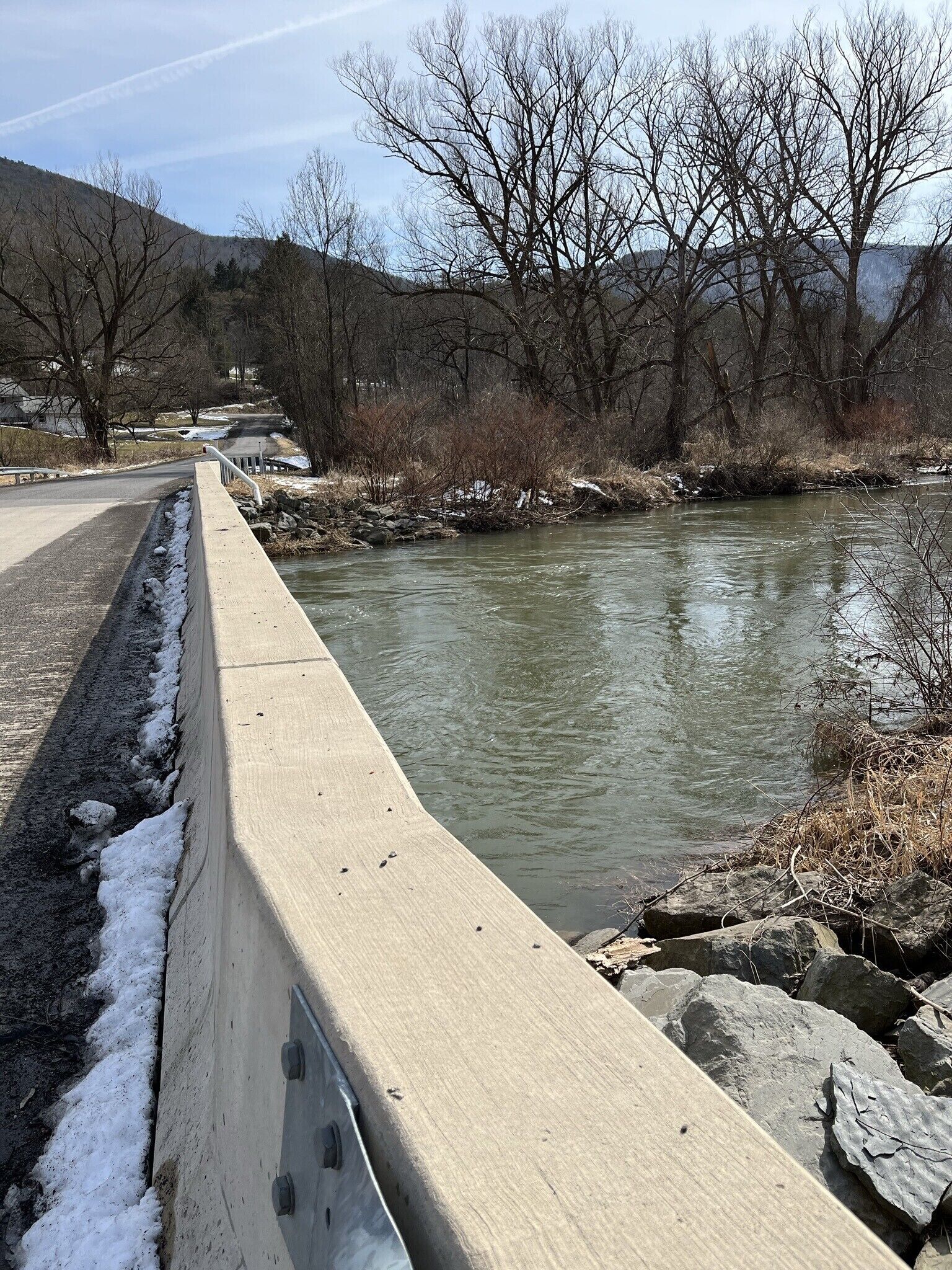 A concrete bridge over a river with bare trees and a hilly landscape in the background.