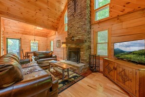 L’intérieur d’une cabane en rondins, avec une cheminée en pierre, un canapé en cuir, un plancher en bois et un téléviseur.
