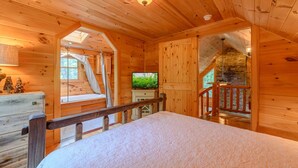 A cozy log cabin bedroom with a bed, a TV, and a staircase leading to a loft area. - Private Creekside Cabin with Hot Tub (Fleetwood)