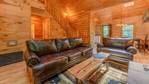 A log cabin interior with a wooden staircase, leather sofas, a dining table, and a chandelier. - Private Creekside Cabin with Hot Tub (Fleetwood)