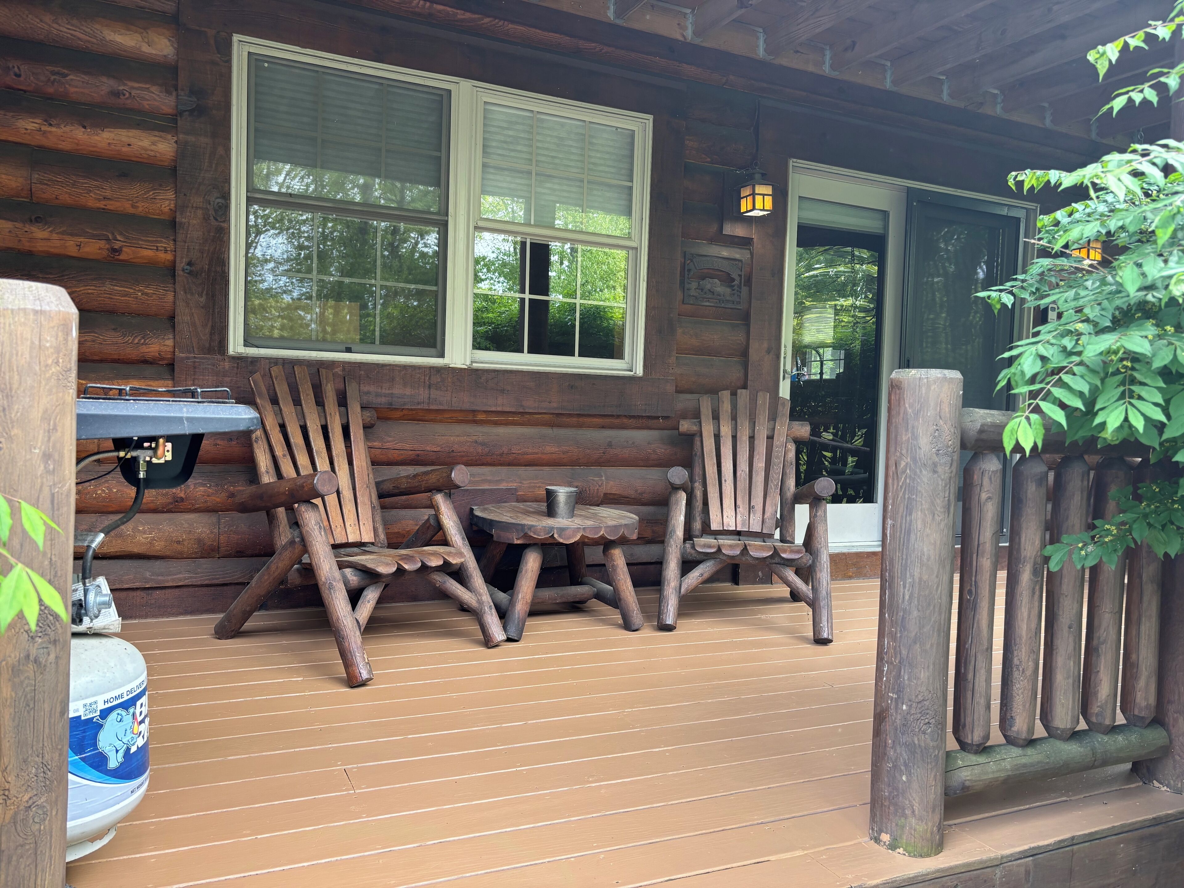 A wooden cabin with a porch, two Adirondack chairs, a small table, and a propane tank.