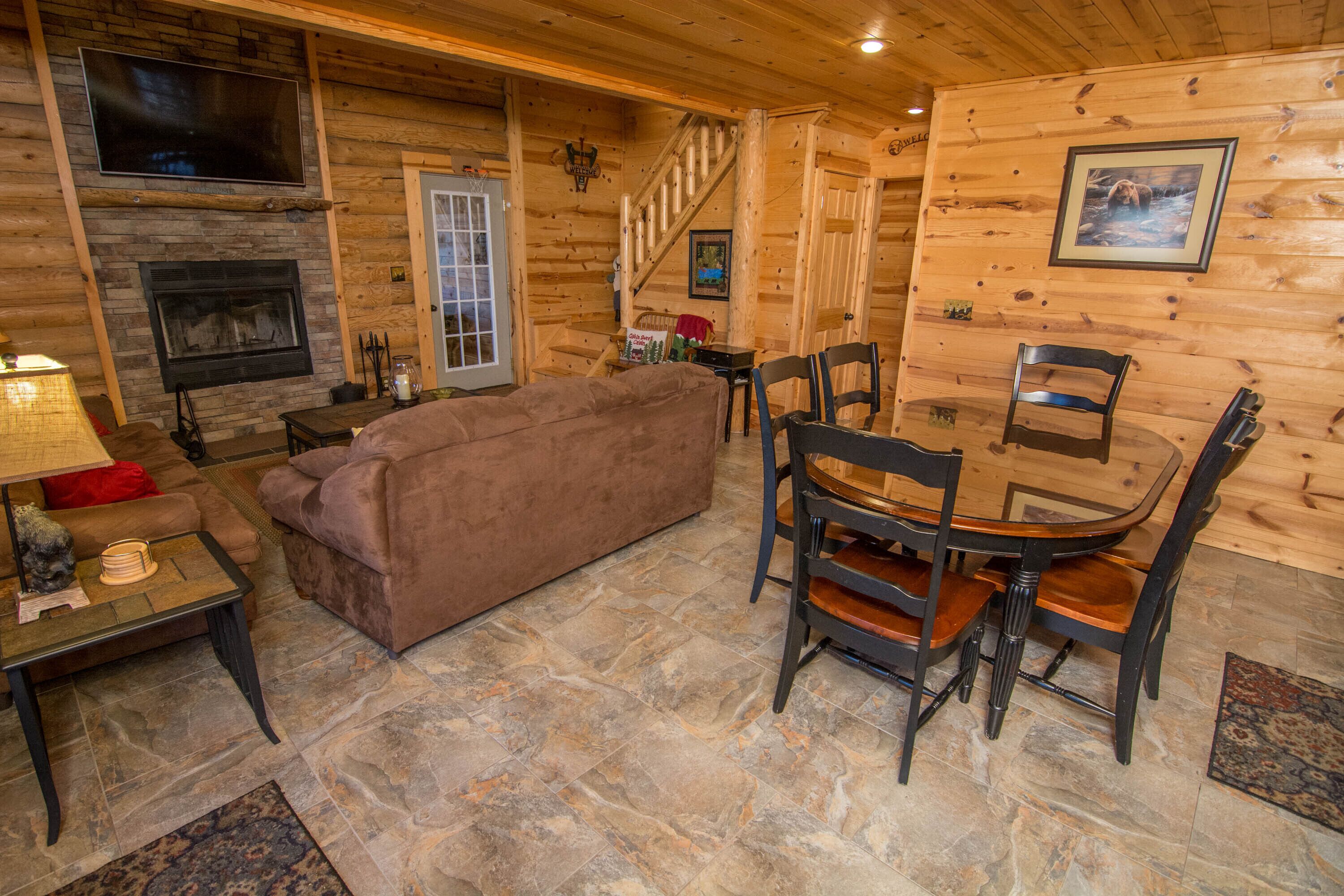 A cozy log cabin interior with a fireplace, a sofa, a dining table with chairs, and a staircase.