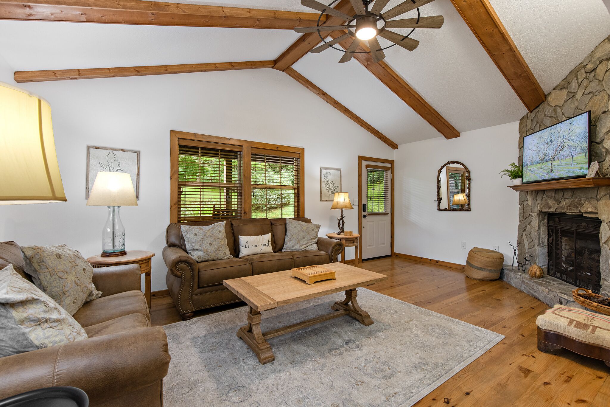 A spacious living room with a wooden ceiling, a stone fireplace, a large window with blinds, and a cozy seating area.