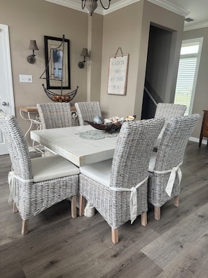 Un coin repas avec une table blanche et des chaises en osier, un tableau encadré, des lampes fixées au mur et un panneau.