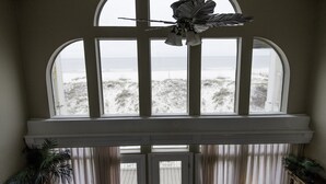 A room with large arched windows overlooking a beach, a ceiling fan, and a view of the ocean.