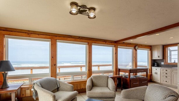 A spacious room with large windows overlooking the ocean, wooden beams, a dining area, and a kitchenette.