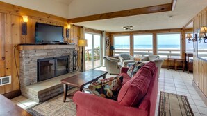 A cozy living room with a stone fireplace, comfortable seating, and large windows offering a view of the ocean.