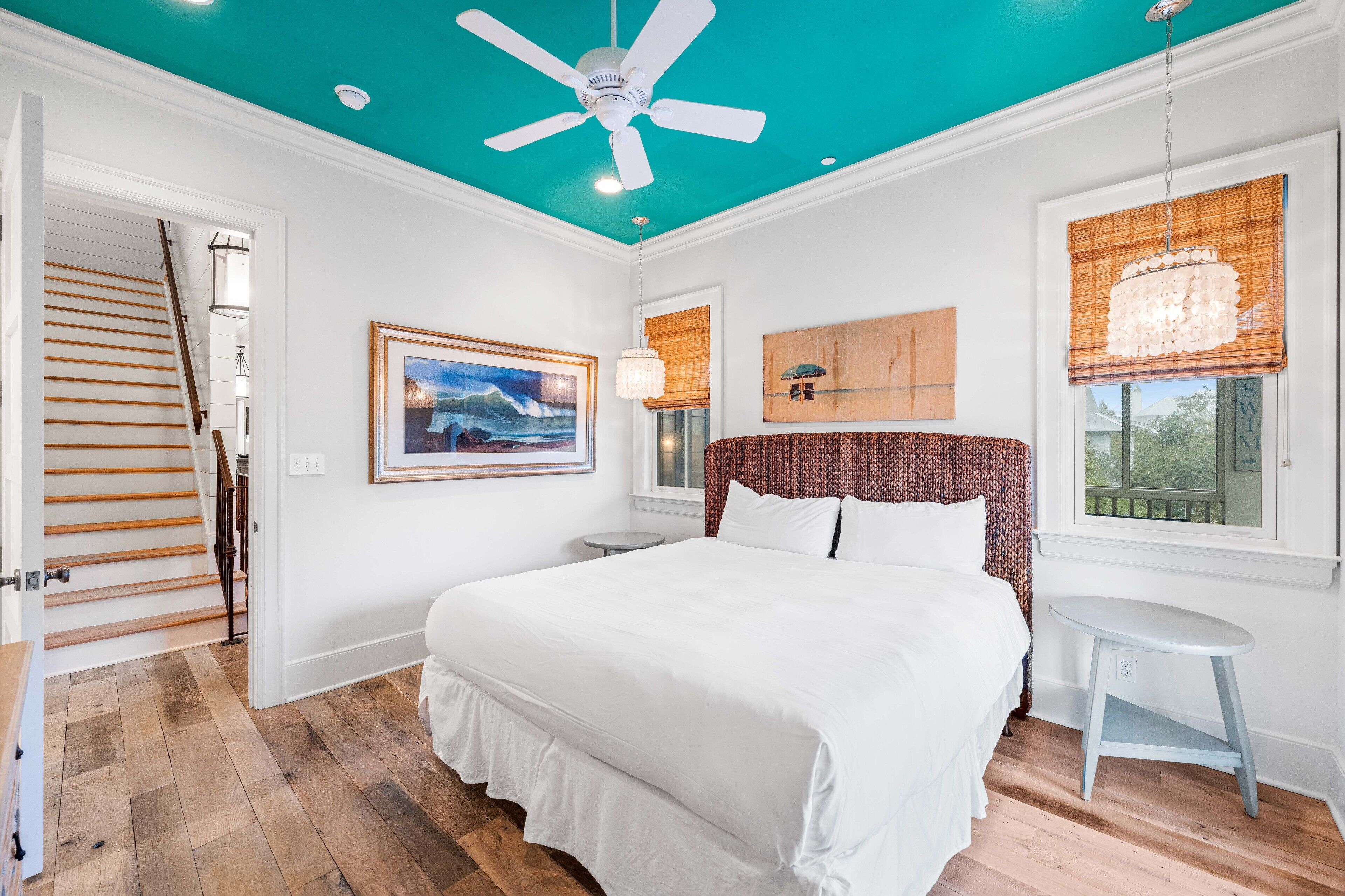 A bedroom with a large bed, a ceiling fan, a wooden staircase, and a view of the outdoors through a window.
