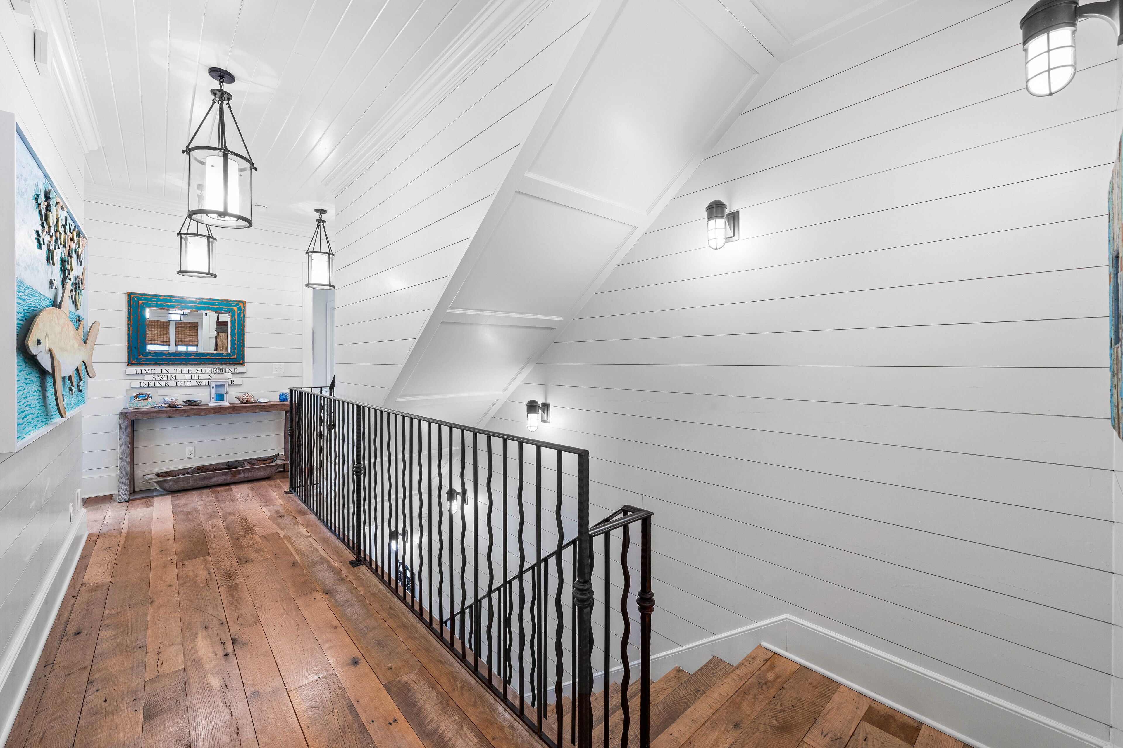 A wooden staircase with a black railing, white paneled walls, and pendant lighting.