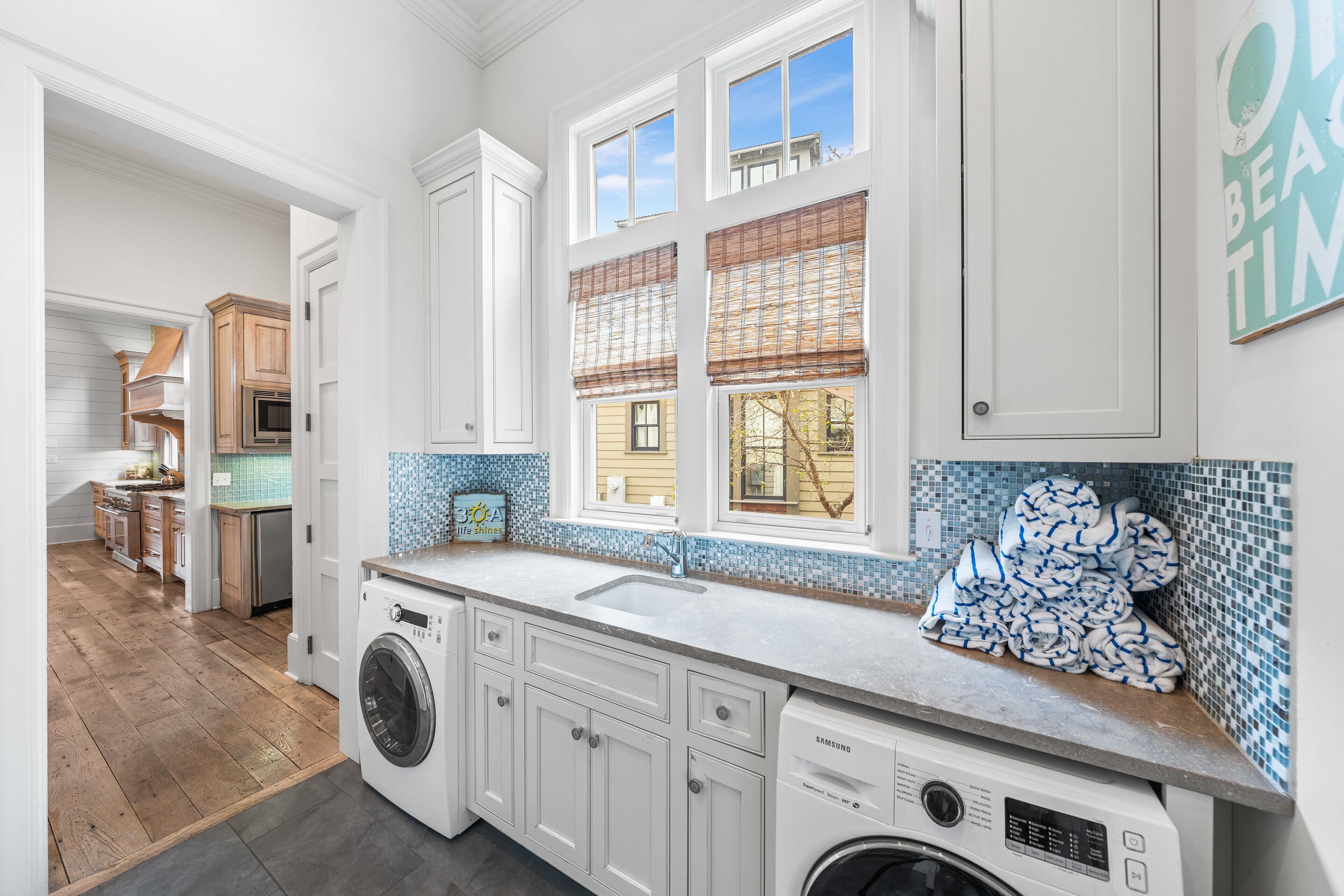 A modern kitchen with white cabinetry, a built-in washer and dryer, a large window with blinds, and a blue-tiled backsplash.