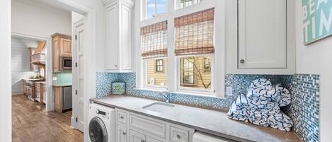 A modern kitchen with white cabinetry, a built-in washer and dryer, a large window with blinds, and a blue-tiled backsplash.