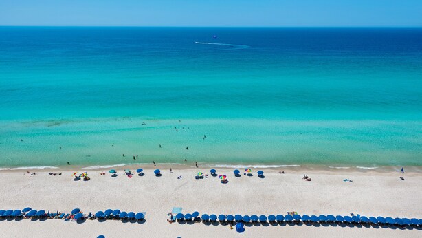 Ein Strand mit klarem türkisblauem Wasser, ein Jet-Ski in der Ferne und Reihen von blauen Sonnenschirmen im Sand.
