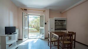 Een kamer met een televisie op een witte standaard, een eettafel met stoelen, en een witte kast met planken.
