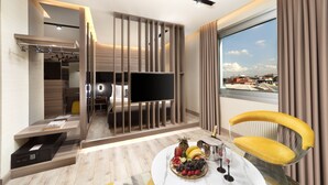 Una habitación de hotel moderna con una televisión de pantalla plana, una mesa de comedor con fruta y vino, una silla amarilla y un ventanal con vistas a la ciudad.