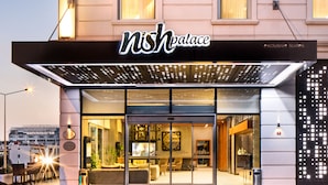 Fachada de hotel de vidrio, con un cartel que dice "nishpalace exclusive suites" y un interior bien iluminado visible a través de las puertas de cristal.