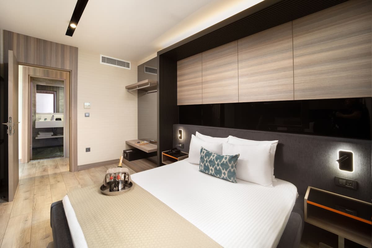 Una habitación de hotel moderna con una cama grande, una mesita de noche, una pequeña nevera y un televisor integrado.