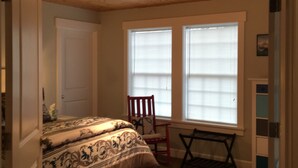 Une chambre à coucher avec un lit, un tapis rouge, un fauteuil à bascule et deux fenêtres équipées de stores.
